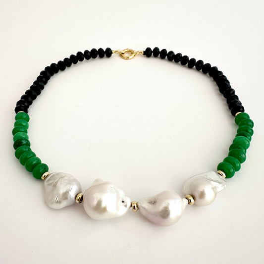 A beaded necklace featuring black round beads, vibrant green faceted beads, and three large white baroque pearls, accented with small gold spacers and a gold clasp, displayed on a clean white background.