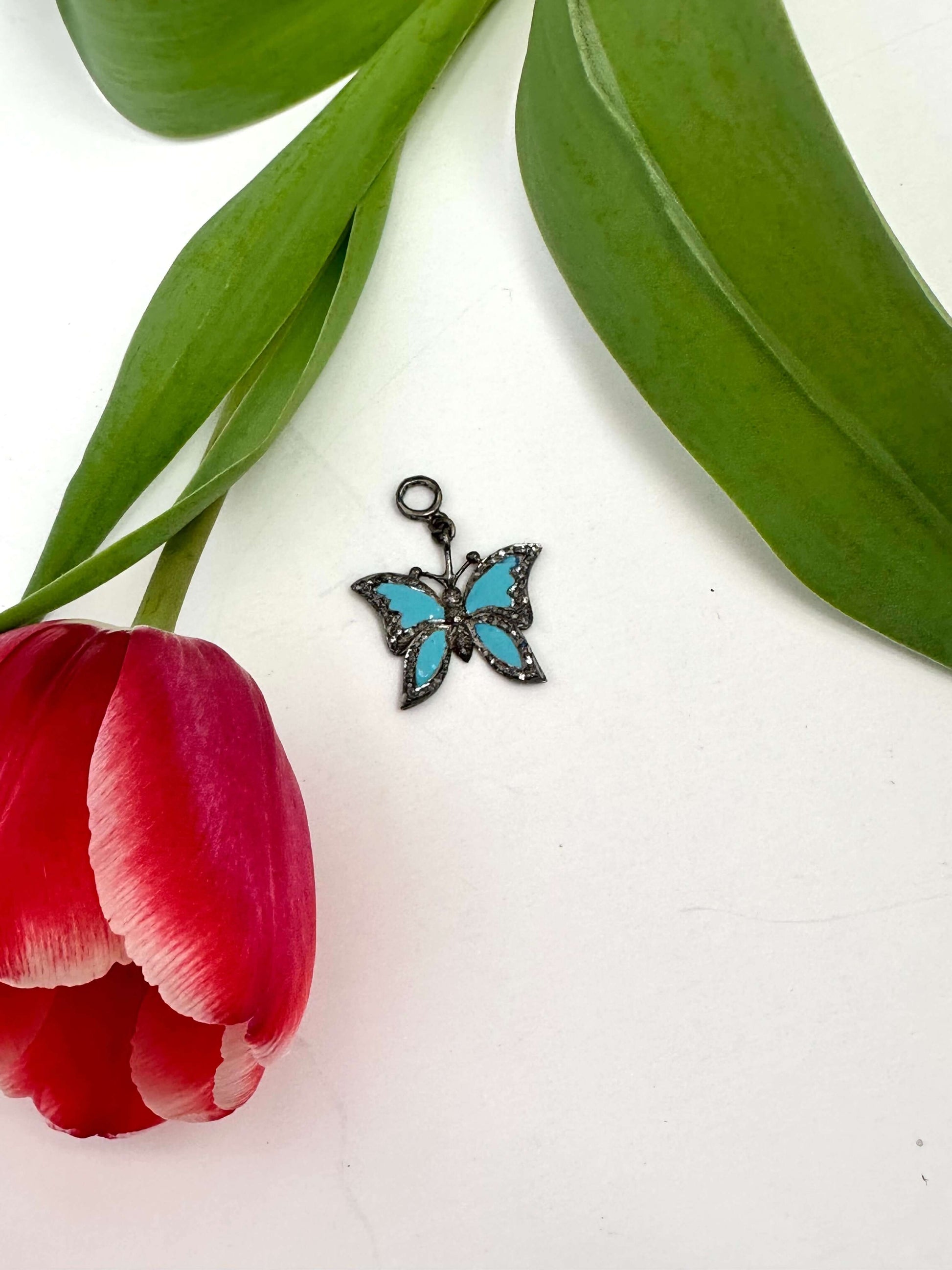 Blue enamel butterfly pendant with diamond pavé displayed next to a red tulip and green leaves.