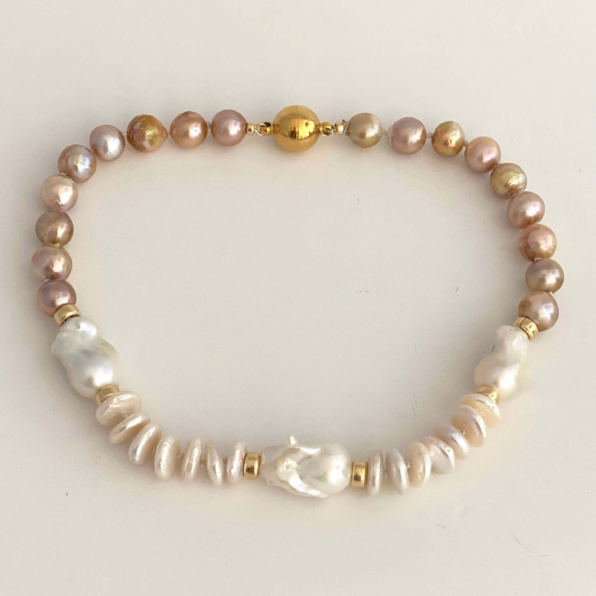 A beaded pearl bracelet featuring a mix of iridescent pinkish-gold, white baroque, and textured white pearls, accented with small gold spacer beads and a central gold bead clasp, displayed on a light cream background.
