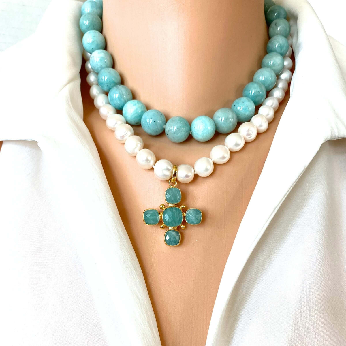 A close-up of a double-strand necklace worn on a mannequin or model's neck, featuring a top layer of light blue round beads and a bottom layer of white faux pearls, with a small gold cross pendant set with light blue stones at the center. The necklace is styled with a white collared shirt.