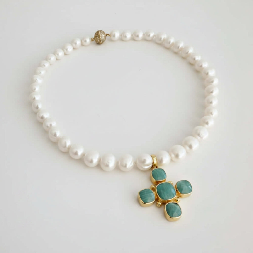 A pearl beaded necklace with a gold cross pendant featuring green cabochon stones, displayed on a light gray background.