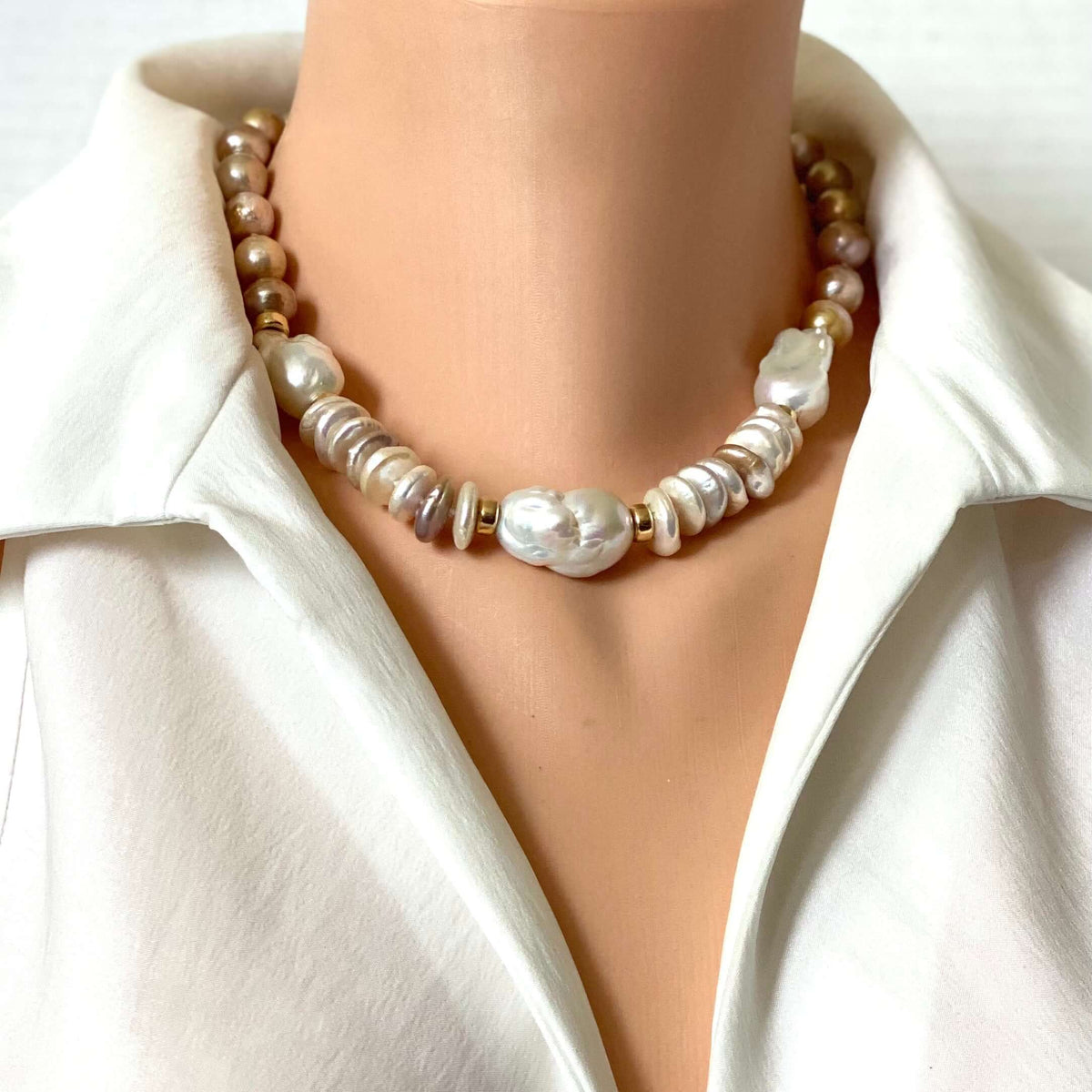 A pearl necklace with mixed white, cream, and beige baroque pearls and small metallic spacer beads, shown on a mannequin torso wearing a white satin shirt.