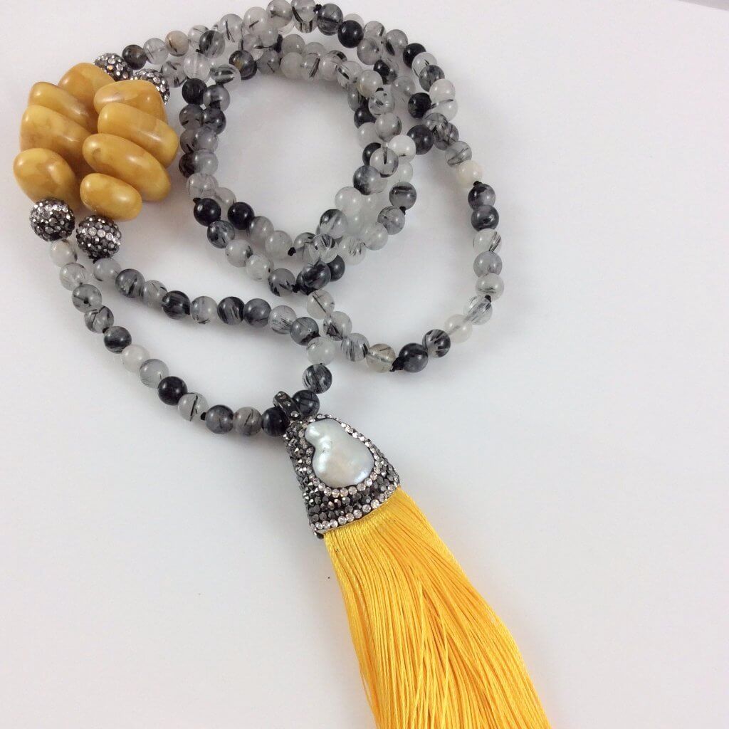 Close-up of rutilated quartz necklace with yellow tassel on white table, hand-knotted beads.