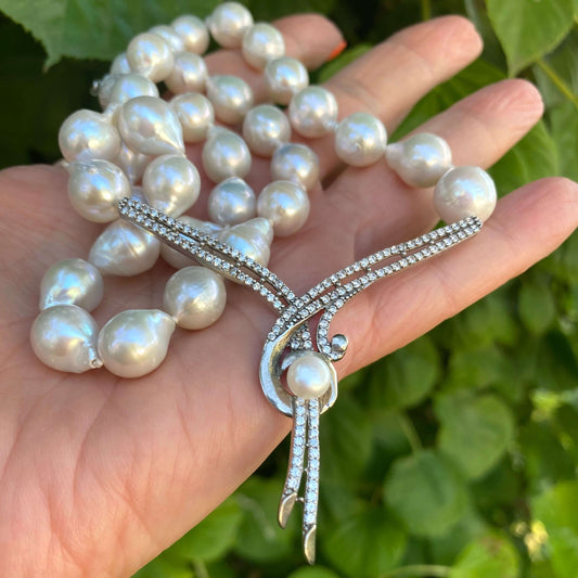 Baroque pearl necklace with sterling silver clasp held in hand against green leaf backdrop, close-up view.