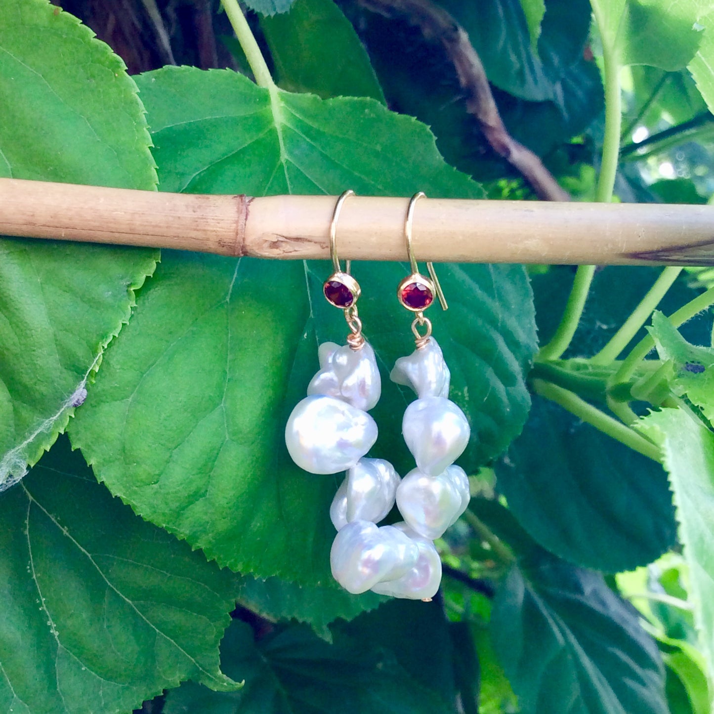 Pearl drop earrings with keshi pearls and red CZ, handcrafted in Canada, showcased against green foliage.