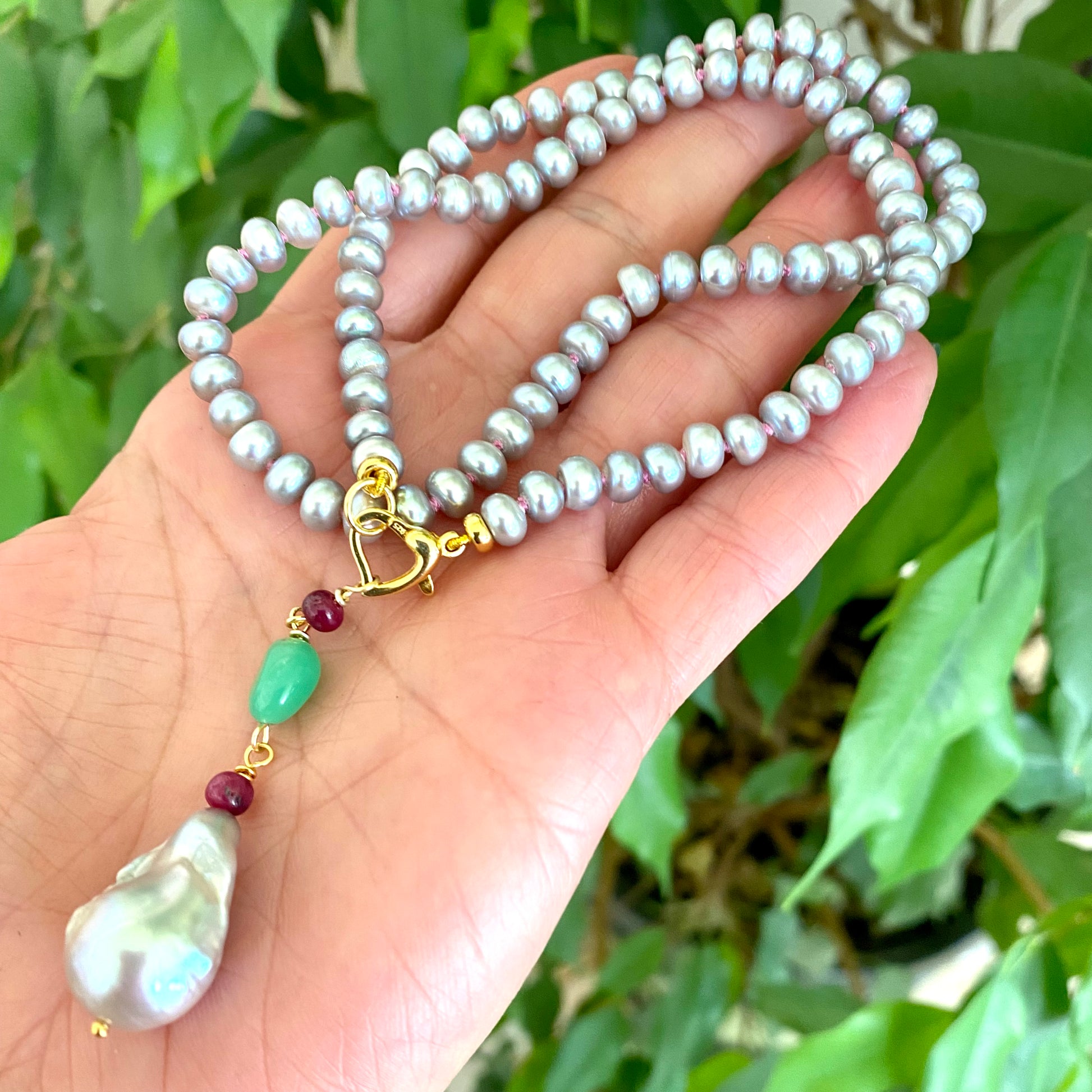 Grey pearl necklace with ruby and chrysoprase pendant displayed in a hand, showcasing elegant freshwater pearls.